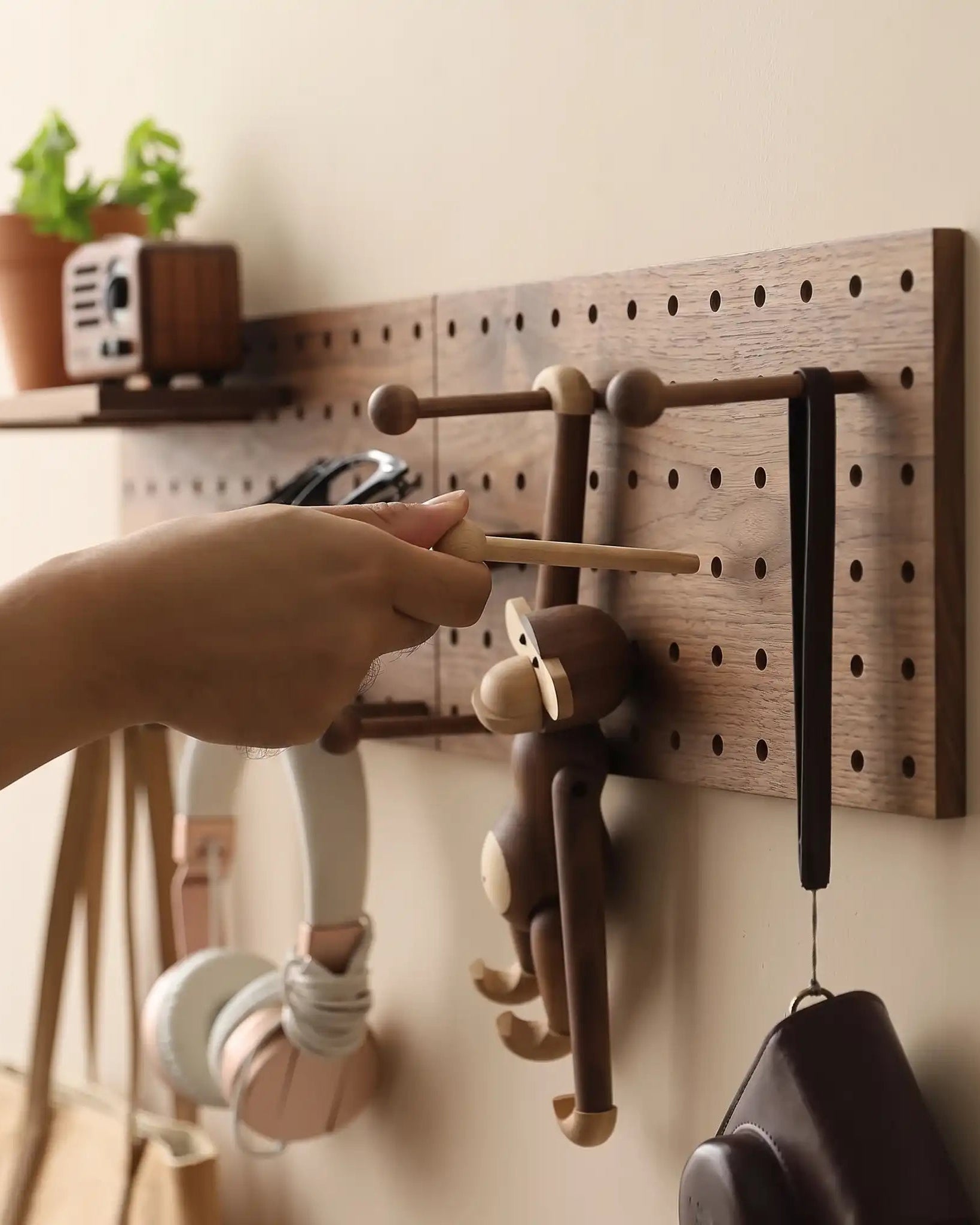 A wooden pegboard wall organizer mounted on a beige wall, with a person's hand adjusting a hanging wooden monkey hook. The rack includes a small shelf with a potted plant and a retro radio, plus hooks holding headphones, a brown bag, and other accessories.