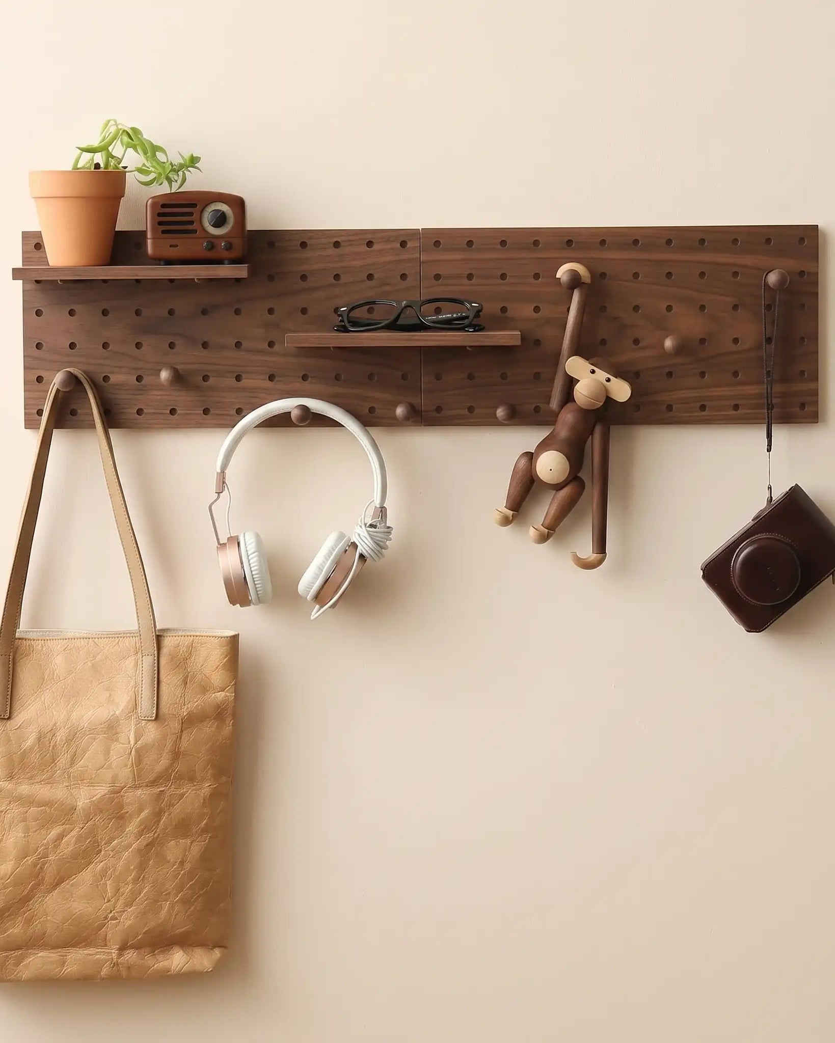 A wooden pegboard wall organizer mounted on a beige wall, with a person's hand adjusting a hanging wooden monkey hook. The rack includes a small shelf with a potted plant and a retro radio, plus hooks holding headphones, a brown bag, and other accessories.